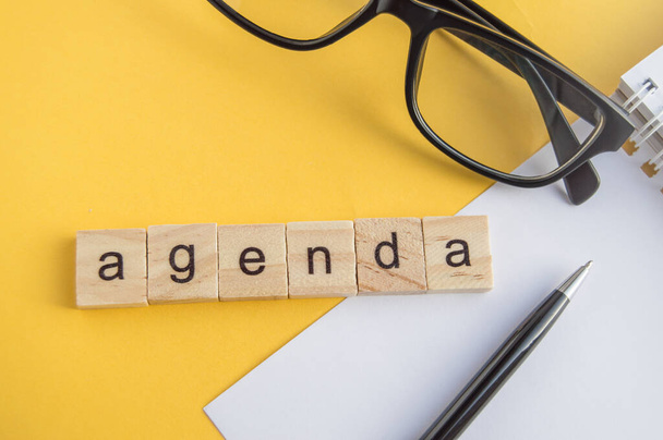 Top view the word of the agenda written on wooden cubes on the desktop with glasses, pen, paper Notepad on a yellow background, top view, flat layout, business style - Photo, Image