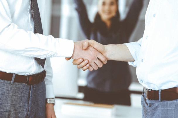 Two businessmen are shaking hands in office, close-up. Happy and excited business woman stands with raising hands at the background. Business people concept - Фото, изображение