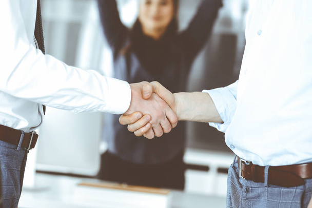 Two businessmen are shaking hands in office, close-up. Happy and excited business woman stands with raising hands at the background. Business people concept - 写真・画像
