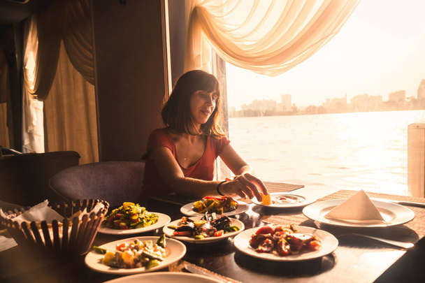 A young tourist having dinner on a boat on the nile a traditional Egyptian meal with sunset light in the window, the city of Cairo in the background. Africa - Photo, image