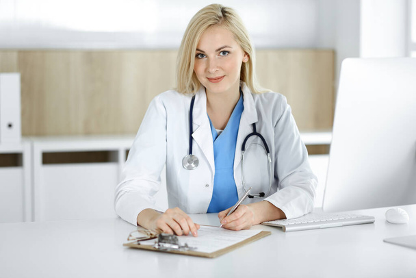 Woman-doctor at work while sitting at the desk in hospital or clinic. Blonde cheerful physician filling up medication history record form - Zdjęcie, obraz