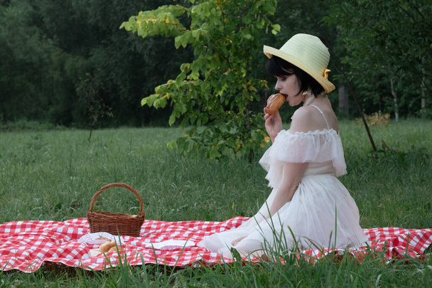 Bella romantica giovane donna con i capelli corti scuri in un cappello di paglia e vestito bianco è seduto su un picnic, in natura - Foto, immagini