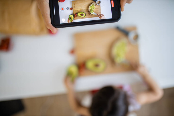 Weibliche Hand mit Smartphone nimmt auf, wie ein kleines Mädchen eine Avocado schneidet - Foto, Bild