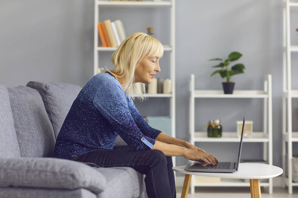 Side view of a mature woman typing on laptop while sitting on her sofa at home. - Foto, imagen