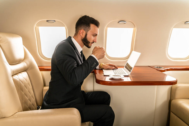 Όμορφος επιχειρηματίας φορώντας κομψό κοστούμι που φέρουν με αποκλειστικό ιδιωτικό τζετ - Επιτυχημένος επιχειρηματίας κάθεται στην αποκλειστική επιχειρηματική θέση στο αεροπλάνο, έννοιες σχετικά με τις επιχειρήσεις και trasportation - Φωτογραφία, εικόνα