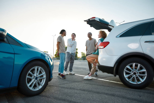 A group of four young casually dressed people speaking to each other smiling and standing together outside on a parking site with cars around - Foto, Bild