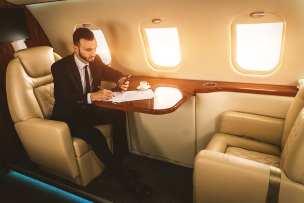 Pohledný podnikatel v elegantním obleku létání na exkluzivním soukromém tryskáči - Úspěšný podnikatel sedí v exkluzivní obchodní třídě v letadle, koncepce podnikání a dopravy - Fotografie, Obrázek
