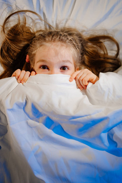 top View. Little girl lies in bed, frightened and covers face with blanket - 写真・画像