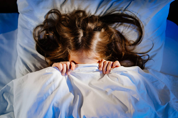 frightened girl covers his face with a blanket in bed in her room. childs fear - Foto, afbeelding