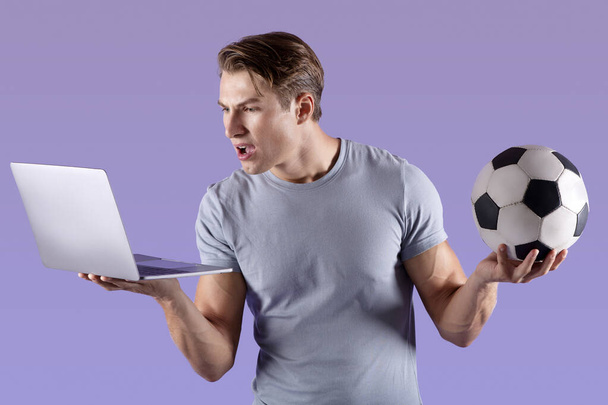 Sportowy człowiek tysiąclecia rozczarowany porażką drużyny piłkarskiej, oglądanie meczu piłki nożnej na laptopie, fioletowe tło studio - Zdjęcie, obraz