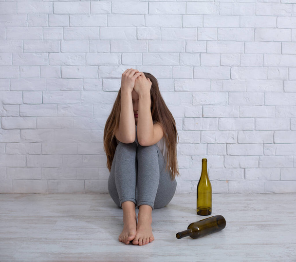 Концепция алкоголизма и депрессии. Молодая женщина плачет на полу дома с пустыми бутылками вокруг нее - Фото, изображение