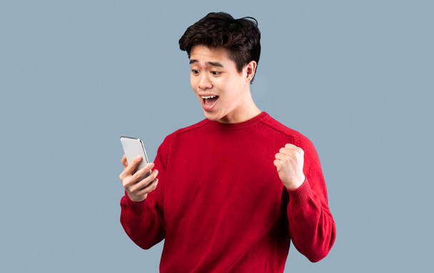Eccitato uomo asiatico utilizzando il telefono cellulare, celebrando vittoria online - Foto, immagini