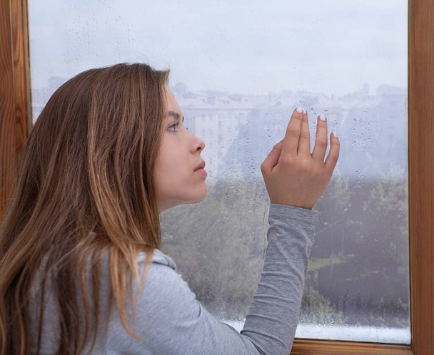 Jovencita solitaria mirando por la ventana, perdida en el pensamiento, tocando el cristal con gotas de lluvia - Foto, imagen