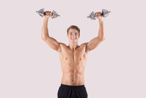 Portret van positieve jonge atleet tillen halters boven zijn hoofd en glimlachen op lichte studio achtergrond - Foto, afbeelding