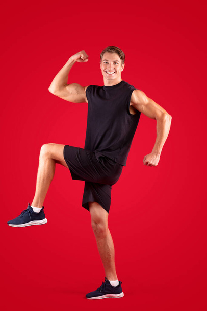 Полнометражный портрет молодого спортсмена, марширующего или идущего с коленями на красном фоне студии - Фото, изображение