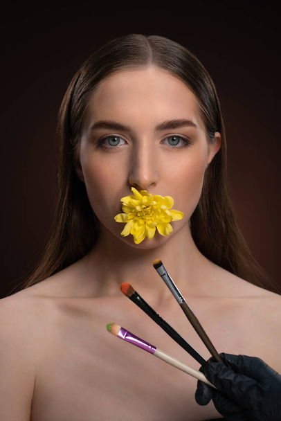 Natural cosmetics concept. Beauty model with bare shoulders holding a flower in her mouth and brushes in front looking at camera isolated on black background. Woman nature cosmetic spa concept - Фото, зображення
