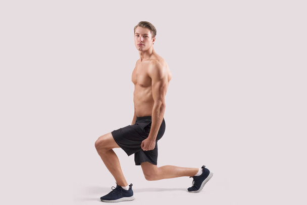 Strong young guy with sexy muscular body doing lunges, warming up before workout over light studio background - Photo, Image