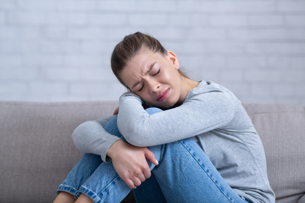 Joven mujer deprimida sentada en el sofá con los ojos cerrados y abrazando sus rodillas, sintiéndose desesperada y estresada - Foto, Imagen