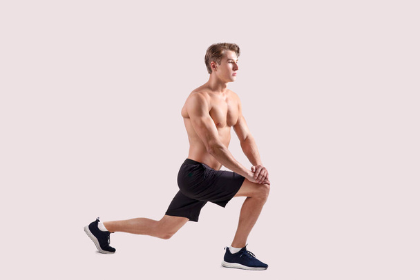 Fit millennial bodybuilder with naked chest doing lunges on light studio background, full length portrait - Photo, Image