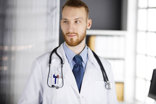 Red-bearded doctor standing straight in clinic near his working place. Portrait of physician. Medicine and healthcare concept - Photo, Image