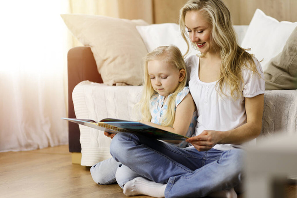 Happy family. Blonde young mother reading a book to her cute daughter while sitting at wooden floor in sunny room. Motherhood concept - Foto, Imagen
