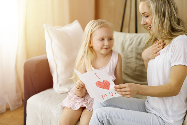 Happy mother day in sunny flat. Child daughter congratulates mom and gives her postcard with heart drawing. Family concept - Фото, зображення