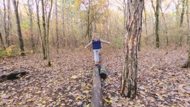 Een meisje in een blauw jasje en een koptelefoon loopt op een boomstam in het herfstbos - Video