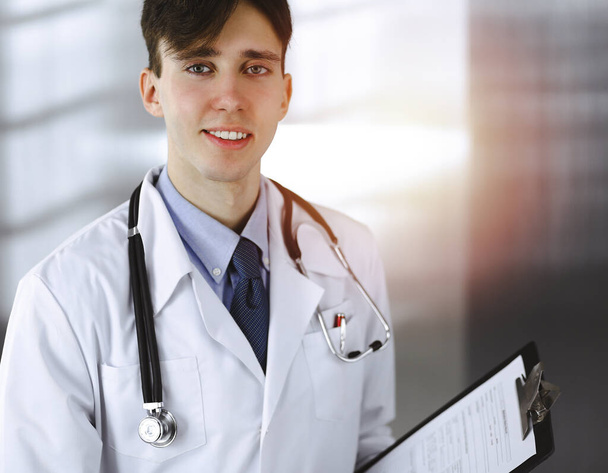 Friendly young male doctor standing with clipboard in modern clinic. Medicine concept - Photo, Image