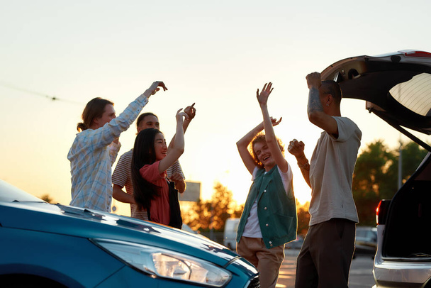 Five young casually dressed friends having a party dancing to music together outside on a parking site near their cars - Foto, Bild