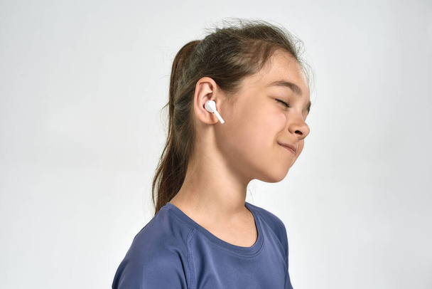 Portrait of little sportive girl child wearing earphones, listening to music, standing with eyes closed while posing isolated over white background - Foto, Imagen