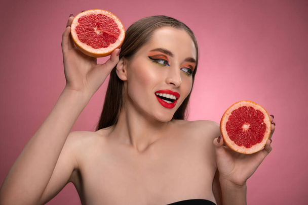 Beautiful young woman model holding two juicy grapefruit sliced in half in her hands looking sideways. Charming joyful funny lady with red lips and long hair isolated on pink background - Zdjęcie, obraz