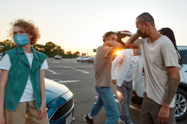 A group of young casually dressed friends of different nationalities greeting each other with elbows wearing masks standing outside on a parking site with their cars on a background - Fotoğraf, Görsel
