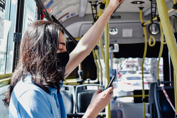 mladý muž kontroluje svůj telefon v autobuse, zatímco cestuje autobusem, proti světlu, s vlasy z klády - Fotografie, Obrázek
