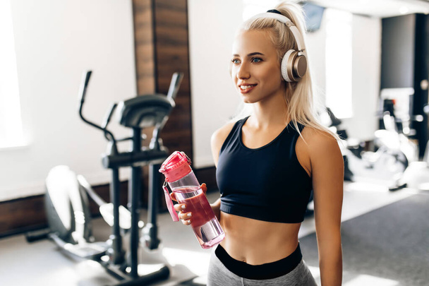 young sportswoman in sportswear and headphones holds a bottle of water, listens to music in the gym - 写真・画像
