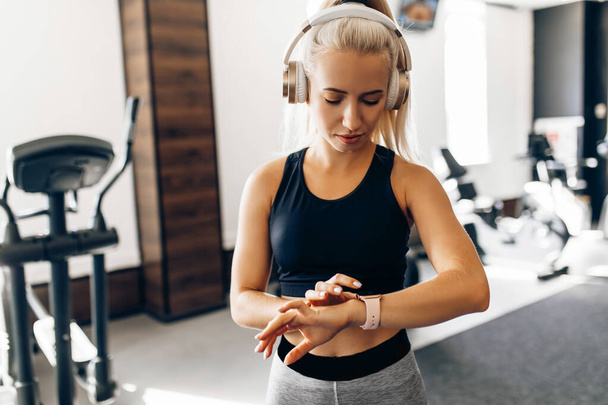Attractive young sportive woman goes in for sports in the gym, listens to music on headphones and looks at the fitness watch on her hand, sports, healthy lifestyle - Fotó, kép