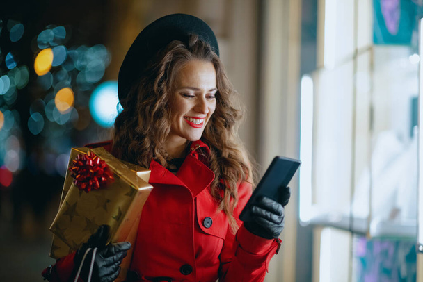 Winter Fun. happy middle aged woman shopper in red coat and black beret with gifts and smartphone buying online outside in the city in the evening. - Photo, Image