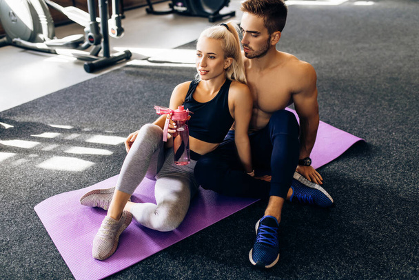young sports couple in sportswear sitting on yoga mats and holding water bottles after workout at the gym - Photo, Image