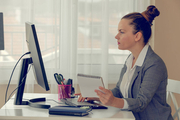 Side view of a girl in white shirt and plaid jacket near her computer in the office. She is looking at the screen and holding a notebook. Online working and education.. Concept of office routine. - Φωτογραφία, εικόνα