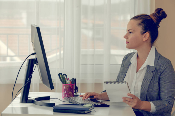 Вид збоку на дівчину в білій сорочці і плетений піджак біля її комп'ютера в офісі. Вона дивиться на екран і тримає блокнот. Інтернет робота та освіта. Концепція офісної рутини
. - Фото, зображення