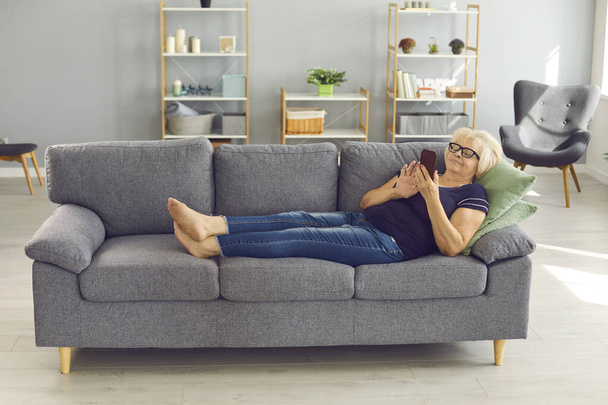 Positive senior woman lying on sofa, relaxing and chatting or making order online on smartphone - Fotoğraf, Görsel