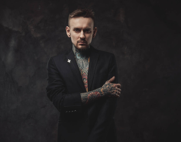 Tattooed serious guy in jacket with stylish haircut in dark background - Фото, изображение