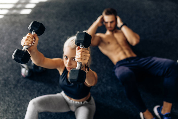 Sportive young couple, man and woman doing abdominal exercises with dumbbells, on the floor in the gym - Foto, imagen
