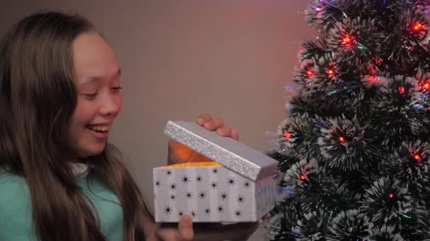 A child girl opens gift from Santa Claus next to Christmas tree. Child opens gift box in light of garland, rejoices and smiles. Family childrens holiday and celebration, winter rest. Merry Christmas. - Footage, Video