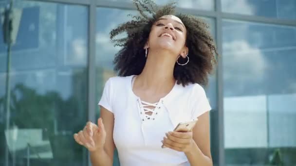 Amerikai afrikai etnikai nő táncol felemeli a kezét a zene gazdaság okostelefon az utcán. Boldog göndör lány vidám érzelmek öröm szereti élvezni az életet és a szabadtéri tevékenységek, lassított felvétel - Felvétel, videó