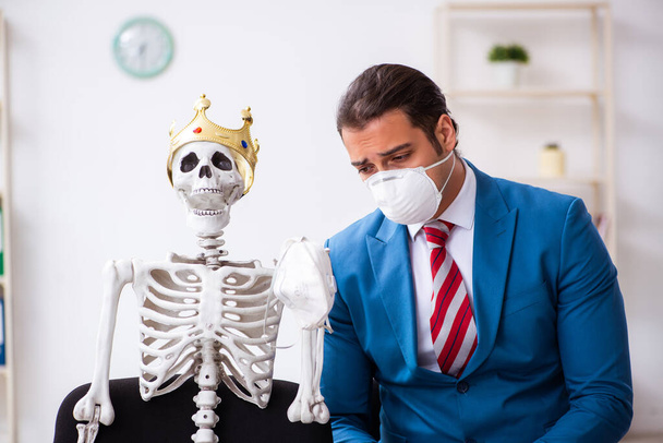 Νεαρός επιχειρηματίας και σκελετός στο γραφείο σε πανδημία έννοια - Φωτογραφία, εικόνα