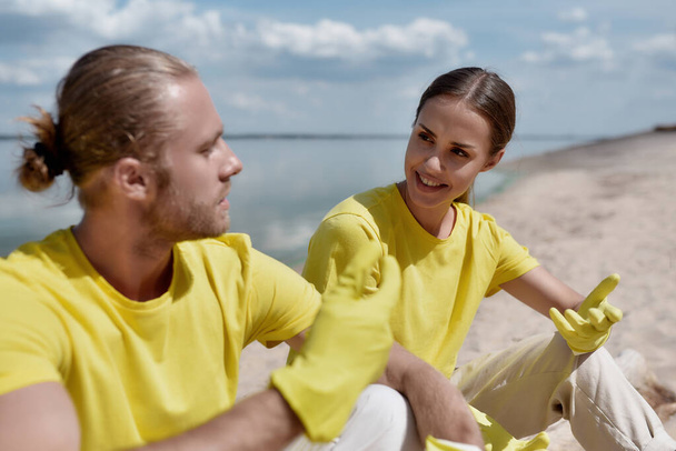 Two young happy eco activists wearing uniform and rubber gloves resting after cleaning a beach and discussing something - Photo, Image