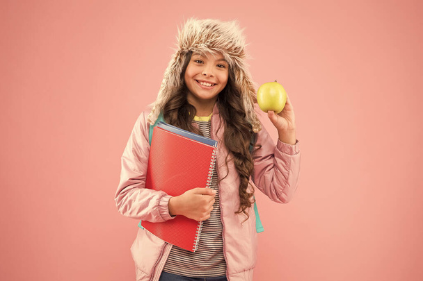 uczennica z książkami po lekcjach. przerwa w szkole. mała szczęśliwa dziewczyna earflap kapelusz jeść jabłko. ferie zimowe. Różowe tło nastolatka. codzienne życie ucznia. Nowoczesna edukacja. Student miesiąca - Zdjęcie, obraz