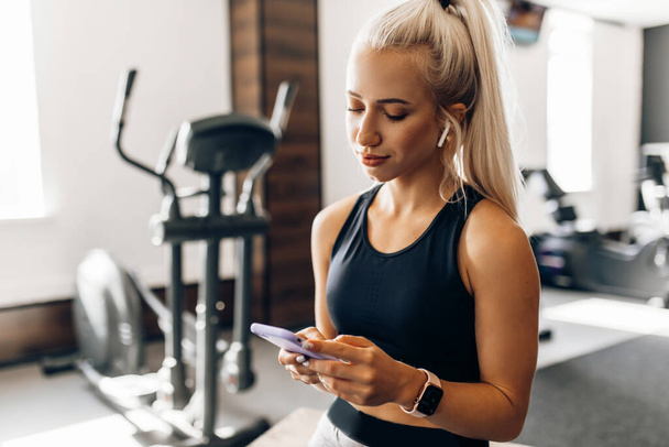 Beautiful young fitness woman, in sportswear, relaxing after a workout uses a mobile phone, in the gym - Fotó, kép