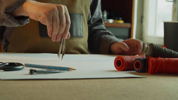 Een kleermaker in een schort maakt een markering met kompassen op het patroon. Op de voorgrond, rollen met gekleurde draden. - Foto, afbeelding