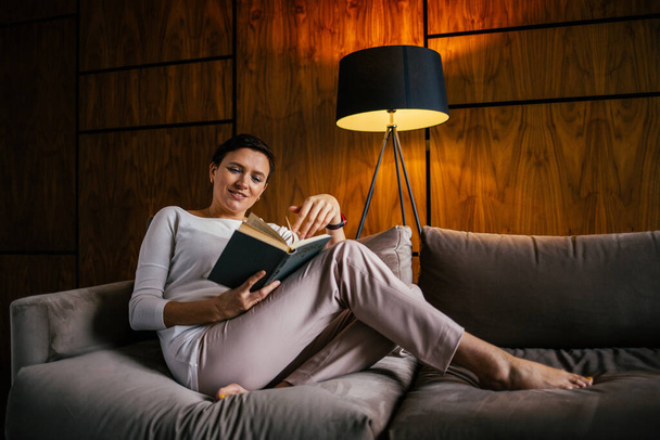 A young woman sits on a sofa in a cozy apartment with a book in her hand - Valokuva, kuva